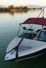 A white boat with a red bimini top docked on calm water, with a black bow sling attached to the front.